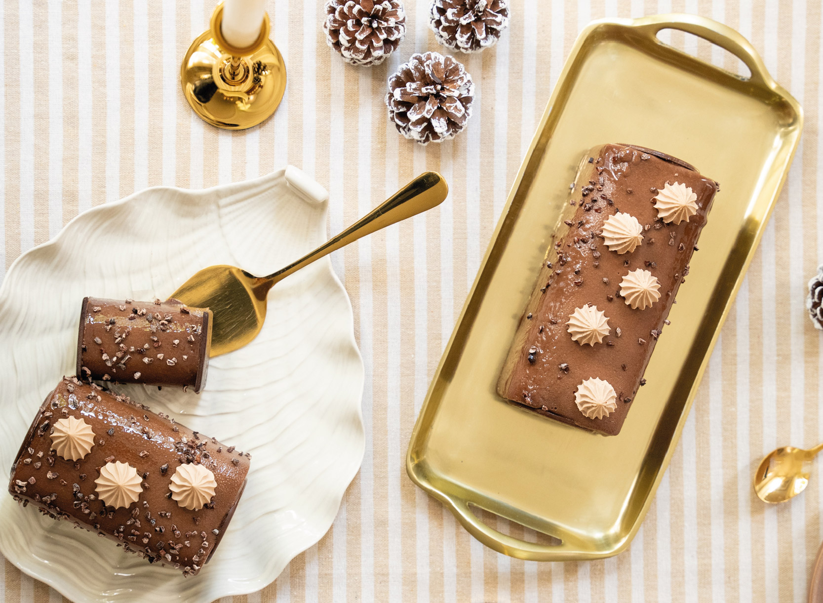 Cette bûche marie la douceur d’une crème brûlée à la vanille, le fondant d’un biscuit chocolat, le croustillant d’un praliné feuillantine et l’intensité d’une mousse au chocolat noir, vue de dessus, elle est déclinée en version une, quatre et six personnes posées sur une nappe rayée blanc et beige avec deux bougeoirs dorés et quelques pommes de pin