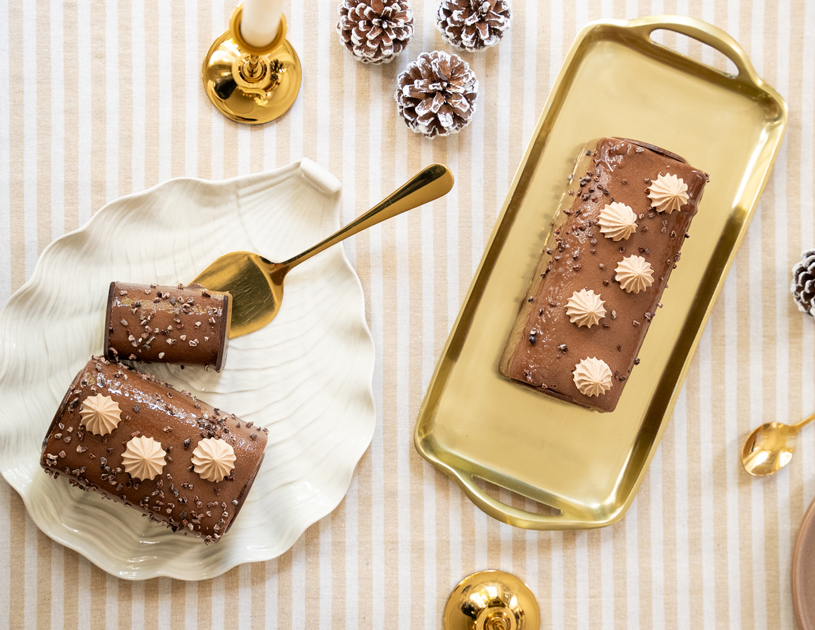 Cette bûche marie la douceur d’une crème brûlée à la vanille, le fondant d’un biscuit chocolat, le croustillant d’un praliné feuillantine et l’intensité d’une mousse au chocolat noir, vue de dessus, elle est déclinée en version une, quatre et six personnes posées sur une nappe rayée blanc et beige avec deux bougeoirs dorés et quelques pommes de pin