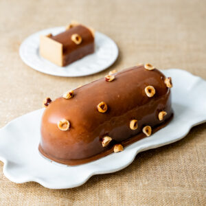 Une bûche au glaçage gianduja brillant enveloppe l’ensemble avec élégance, rehaussé par des éclats de noisettes torréfiées.