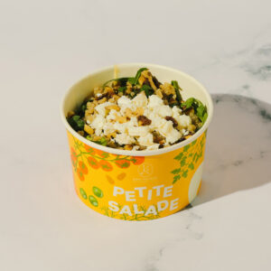 Une petite salade faite de roquette, trio de quinoa, menthe fraîche, fenouil, chèvre frais et noix dans un bol en carton sur un fond de marbre blanc